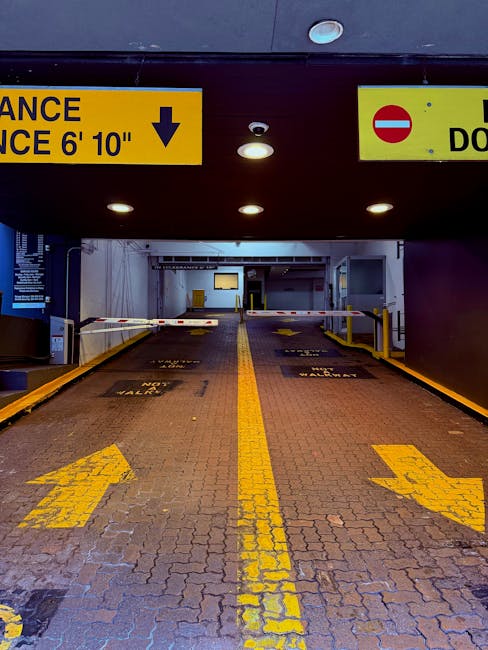The image shows the entrance to an underground parking garage with a height restriction sign indicating 6 feet 10 inches. The foreground features a brick-paved driveway with yellow directional arrows and parking space markings, leading towards the garage's internal area. Barriers and retractable gates are visible at the entrance, with parking lane markings and safety cones beside the driveway. Overhead lighting illuminates the entrance area, and yellow and black safety bollards are positioned along the sides of the driveway. Inside the garage, various structural elements, including walls, signage, and additional barriers, are visible, indicating a controlled entry for vehicle access. This setting aligns with aspects of moving logistics, such as transporting furniture and packing materials during home relocation, which Mayfair Man and Van handles as part of their removals services.