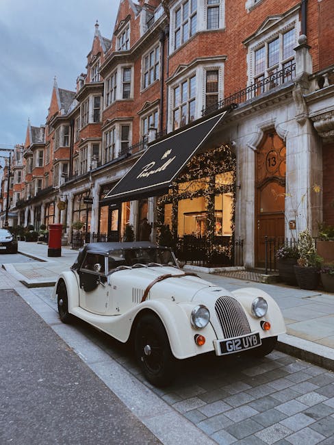 Dusk lighting illuminates a row of elegant, white Victorian-style townhouses in Mayfair, featuring ornate columns, black wrought-iron balconies, and large sash windows. The pavement in front is lined with a series of parked vehicles, including a silver estate car, a black sedan, and other cars, situated on designated parking bays along the street. A black van belonging to Mayfair Man and Van is partially visible, positioned close to the curb, presumably involved in a home relocation process. The image shows clear evidence of furniture and boxes being loaded or unloaded, with a person in dark clothing visible near the entrance of one townhouse. Surrounding the scene, there are lush green trees extending branches over the street, and Union Jack flags are displayed on flagpoles attached to some of the buildings, contributing to the classic London ambiance. This scene encapsulates the typical environment where professional removals and packing services operate within Mayfair's upscale residential area, integrating the logistics of furniture transport and house moves.