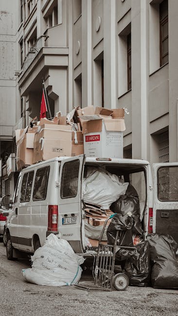 A white panel van parked on a city street in front of a multi-storey building with a beige facade and window balconies. The van’s rear doors are wide open, revealing a variety of packed moving boxes