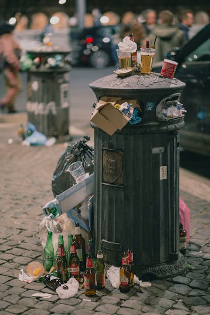 Three large plastic waste bins with ribbed surfaces, two yellow and one green, are positioned outside a residential building on a paved area. The yellow bins are placed side by side, with the green bi