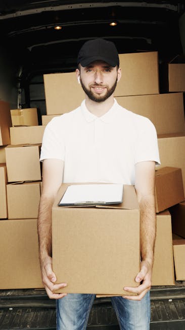 A man wearing a black cap, white t-shirt, and earphones stands inside the open rear of a van used for house removals. He is holding a cardboard box with both hands, preparing for a furniture transport