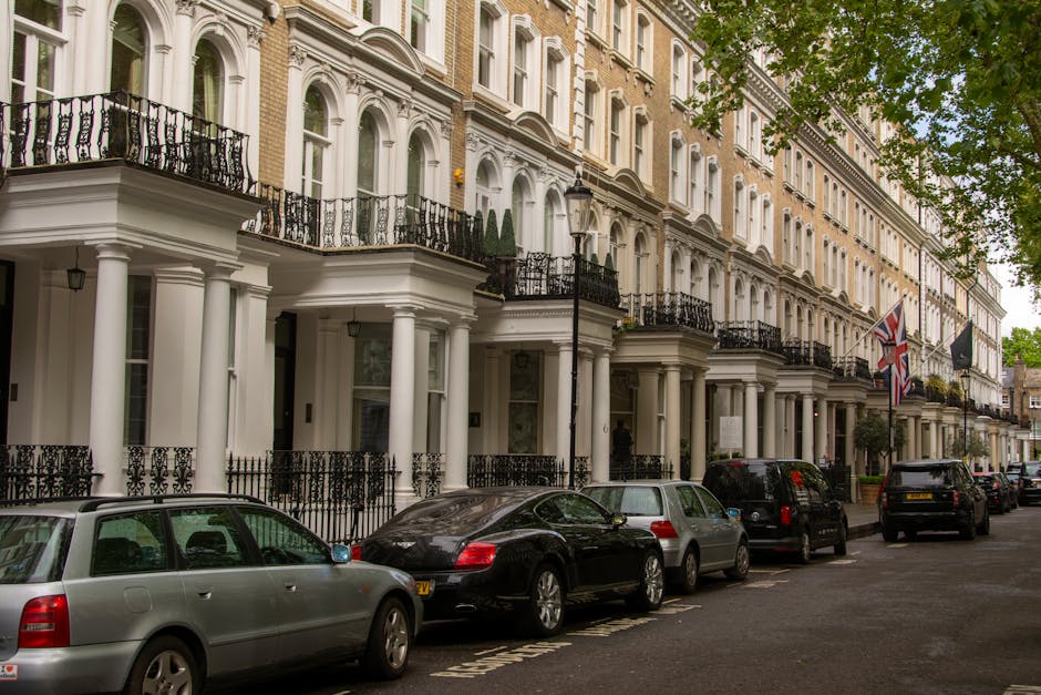 The image shows a row of elegant, multi-storey townhouse-style buildings with white facades, decorative columns, and black wrought-iron balconies, located on a residential street. Several cars, including a silver estate, a black luxury vehicle, and other sedans, are parked along the pavement, indicating a typical urban setting in a high-end area such as Mayfair. Green foliage from trees partially shades the street scene, with some leaves visible at the top right corner. The buildings display classic architectural features, including large windows, ornate moldings, and flags mounted on poles near the entrances. The street scene suggests a quiet, upscale neighbourhood suitable for home relocations, with professional removals services like Mayfair Man and Van handling furniture transport and packing activities. The image captures a moment of vehicle loading or unloading outside the residential properties, reflecting the logistics involved in house removals and furniture transport in luxury apartments like those on Park Lane Flats.