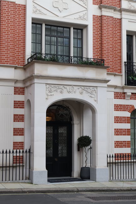 The image shows the exterior of a historic brick building with white stone detailing around the entrance, including an ornate archway and decorative carvings above the doorway. A black door with glass panels and metal fittings is set within the arch, flanked by two small, black metal fences with pointed finials along the pavement. To the right of the entrance, a potted topiary tree with a rounded canopy is placed in a grey container. The upper level features large windows with black frames, and a small balcony with a decorative black wrought iron railing and potted plants. The surrounding environment suggests a residential area suitable for house removals and professional furniture transport, with Mayfair Man and Van providing relocation services for luxury apartments such as Park Lane Flats.