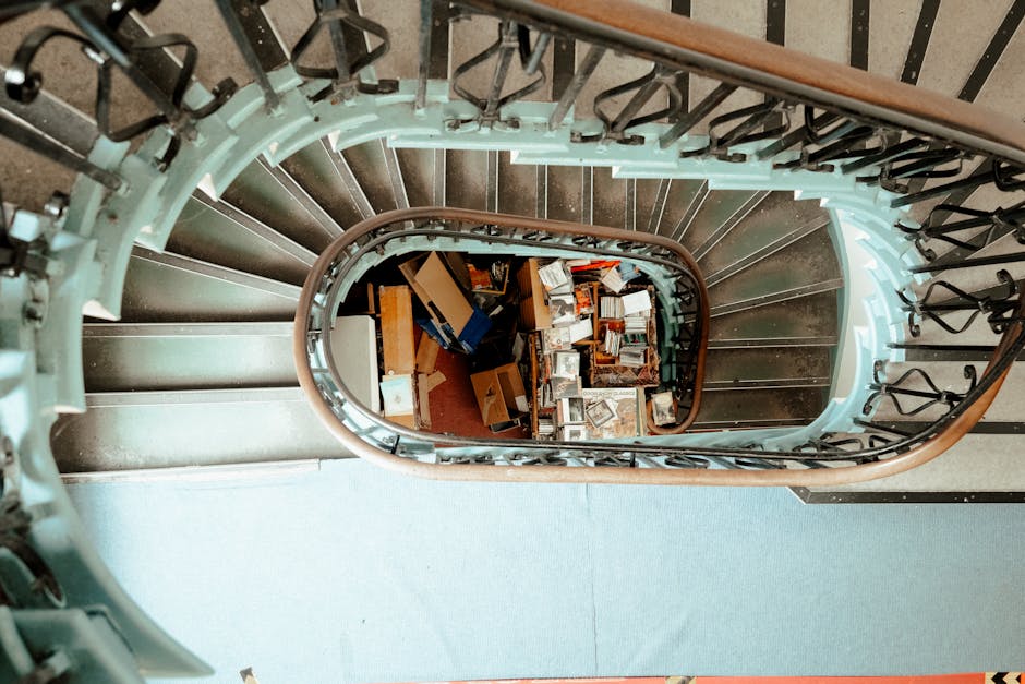 A narrow wooden staircase with dark, polished wooden steps and a curved wooden handrail is shown from an overhead perspective inside a property. The staircase, illuminated by natural light from an uns